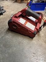 Red Warn PullzAll portable electric winch placed on concrete floor, showing side with product labels and handle.