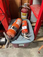 Three fire extinguishers, two orange and one red, shown on and near a plastic storage bin.