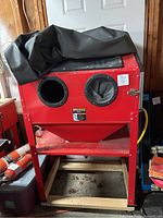 Red powder coated steel floor blast cabinet with viewing window, integrated rubber glove ports, on metal legs atop wooden pallet