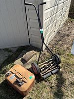 Photo showing Great States manual push reel lawn mower with black frame and rust on cutting reel, positioned next to a rust-colored metal fuel tank on grass beside a building.