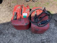 Photo of two red metal gas cans on dry ground, one with hoses attached and visible wear.