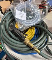 Photo of multiple coiled hoses including green air hose with brass nozzle and trigger handle, yellow and blue hoses coiled underneath and clear braided PVC tubing in sealed packaging.