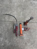 Photo shows one red E Grip grease pump with manual lever and flexible hose, and three hydraulic jacks of various types and sizes on concrete floor.