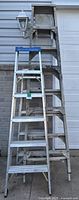 Three aluminum step ladders of different heights leaning against a wall: 5ft, 6ft, and 8ft sizes. Blue caps on top and weathered surfaces visible.