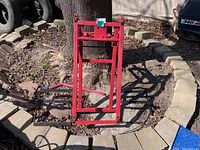 Red metal lawn mower lift shown fully assembled and folded, positioned outdoors on stone and soil ground beside a tree.