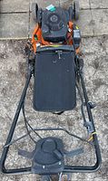 Top-down view showing fabric grass catcher bag attached to the rear of the orange lawn mower deck and black metal handlebar with controls.