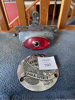 Photo showing the motorcycle tail light with metal mounting bracket and red lens, attached to a metal backing plate, along with the Eagle Spirit derby cover with engraved eagle and inscriptions.