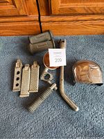 All motorcycle parts displayed on carpet, including foot pegs, kickstand, and horn cover