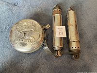 Photo of two chrome motorcycle shocks showing wear and surface rust beside a round chrome air cleaner cover with embossed text and design.