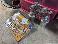 Two metal paint spray guns each with attached metal paint containers, placed on concrete floor next to a sealed air accessory kit in cardboard packaging.