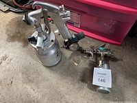 Two pneumatic paint sprayers visible on the floor, showing the full guns and attached paint cups, one with top-mounted cup and one with bottom-mounted larger cup with regulator.