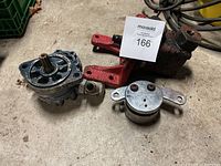 Three mechanical parts placed on concrete floor including a hydraulic pump with red mounting arms and two other metallic components.