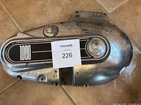 Front and side views of a metal motorcycle primary cover with black and white 1000 emblem, fuel cap, and circular window, showing surface wear.