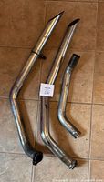 Three metal vehicle exhaust pipes arranged on floor, showing size relative to meter stick, with visible chrome finish and angled outlets