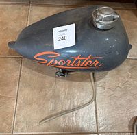 Gray motorcycle gas tank with orange 'Sportster' lettering, silver fuel cap on top, fuel line attached on underside, shown on tiled floor.