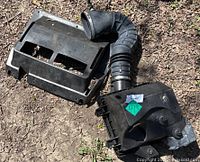 Photo of vehicle air intake system with airbox and flexible hose connected, lying on dirt ground.