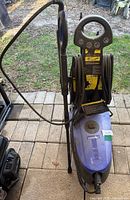Full profile of blue Simoniz pressure washer with spray wand and hose coiled, showing dirt and outdoor use.