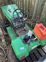 Side and rear view of green Rally riding lawn mower with red gas container on rear fender, black steering wheel, and gear shift exposed. The mower is placed outdoors on pallets and surrounded by fence and foliage.