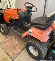 Side angle view showing the orange body of Husqvarna LT125 riding lawnmower, black steering wheel and seat, tires, and part of the engine compartment.