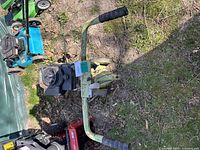 Top view showing the handlebars, engine, and wheels of the rototiller on ground with grass and leaves.