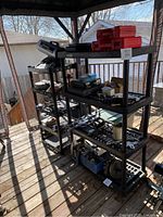 Photo of two black plastic shelving units with various items on them, located outdoors on a wood deck, shown to represent the shelves to be sold.