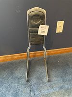 Harley Davidson sissy bar backrest with embossed logo, mounted on chrome frame, standing upright on blue carpeted floor against blue wall with white baseboard and electrical outlet visible.