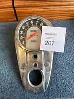 Motorcycle speedometer and dash assembly with odometer reading zero, chrome metal dash panel with GEN and OIL indicator light windows, placed on a blue carpet.