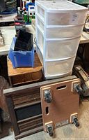 Two plastic 4-drawer storage bins, blue organizer bin, antique wooden scoreboard, furniture dolly, and wood corner shelf visible on table and floor.