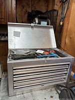 Front angled view of silver metal tool chest sitting on a concrete floor, lid open, showing interior top compartment. Four horizontal drawers visible. Black side handle seen. Surrounded by workshop environment.