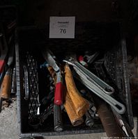 Various hand tools inside a black plastic crate, including machete, adjustable wrenches, ratchet wrench, and hand tools with wooden handles.