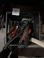 Photo showing a black crate filled with assorted hand tools including hammers, wrenches, and other metal tools with wooden and taped handles.