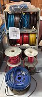Overall view of assorted electric wire rolls on metal rack and spools on floor.