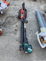 Photo of two gas-powered yard tools lying on concrete, including a red and black Craftsman blower and a green Yardworks curved shaft trimmer.
