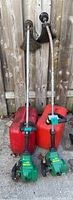Two Feather Lite gas weed whackers leaning against fence with two red gas cans at base