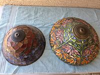 Two Tiffany style stained glass lampshades placed on a blue towel, showing one with floral pattern and the other with geometric design.