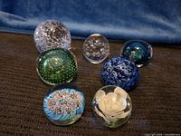 Seven glass paperweights arranged on dark fabric surface with blue backdrop. Varied sizes and designs visible.