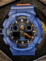 Close-up of blue Casio G-Shock watch face with digital and analog time display, orange and white markings, labeled G-SHOCK and PROTECTION