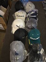 Lot of 11 baseball-style caps arranged on a bed, featuring Puma, TaylorMade, and a green TaylorMade hat. Mostly new with some in plastic wrapping.
