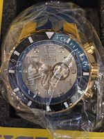 Close-up of the men's Invicta chronograph watch face wrapped in protective plastic showing gold-tone bezel, black dial outer ring, and silver dial with subdials and markings.