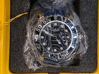 Close-up image of the men's Invicta wristwatch in original plastic wrap, showing black dial with white markers and date window, blue strap, and black bezel with large numerals.