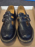 Front view of black Dr. Martens leather shoes with double buckle straps and perforated design on toe area, showing yellow stitching on sole edge.