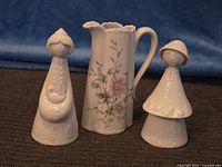 View of three porcelain items: floral pitcher flanked by two white figurines on a brown surface with blue backdrop
