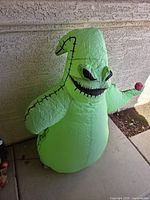 Front view of light green inflatable Oogie Boogie figure with black stitch detailing and red ladybug in hand.