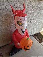 Inflatable character sitting against a textured wall, showing detail of face, horns and pumpkin held in arms.