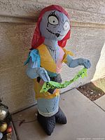 Front angle photo of inflatable Sally figure, holding a green sign with text, standing upright on concrete floor near a wall.