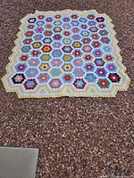 Full view of the quilt laid out on gravel ground showing colorful floral hexagonal patterns and scalloped edges.