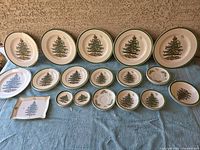 Wide view of the entire Spode Christmas dinnerware set arranged on a blue cloth, showing all plates, bowls, and serving pieces with Christmas tree motif and green rims.