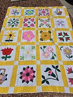 Top view of full quilt with various floral patchwork blocks separated by yellow sashing and white cornerstones, laid on gravel outdoors.