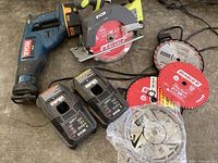 Ryobi cordless reciprocating saw with battery, Ryobi circular saw, two battery chargers, and multiple extra circular saw blades laid out on floor.