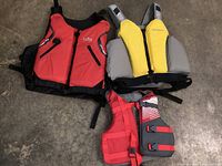 Three life jackets laid on a concrete floor: red Gill adult, gray and yellow adult, and red and gray youth.